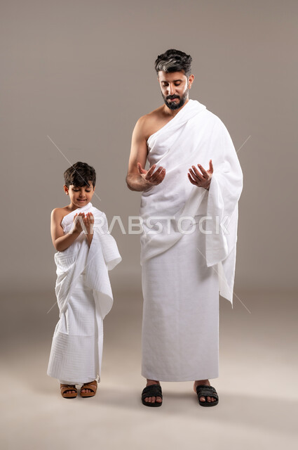 إيماءات التمني طلب المغفره وتحقيق الامنيات, العبادة والتقرب إلى الله بأداء فريضه الحج والعمره بورتريه لرجل وصبي عربيان خليجيان سعوديان يرتديان ثياب الاحرام يرفعان يديهما للاعلى تضرعا الى الله في الحرم المكي الشريف، الاجتهاد في الطاعات والعبادات، خلفيه رماديه