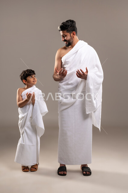 إيماءات التمني طلب المغفره وتحقيق الامنيات, العبادة والتقرب إلى الله بأداء فريضه الحج والعمره بورتريه لرجل وصبي عربيان خليجيان سعوديان يرتديان ثياب الاحرام يرفعان يديهما للاعلى تضرعا الى الله في الحرم المكي الشريف، الاجتهاد في الطاعات والعبادات، خلفيه رماديه