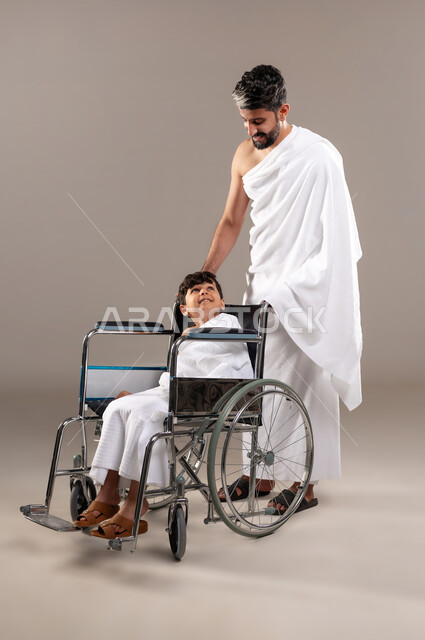 الاستعداد لاداء مناسك الحج والعمره والتقرب الى الله بالاعمال الصالحه، بورتريه لرجل عربي خليجي سعودي يرتدي ثوب الاحرام يجر ابنه الصبي المقعد من ذوي الاحتياجات الخاصه لاداء مناسك الحج او العمره، فريضه الحج هو الركن الخامس من أركان الاسلام لمن استطاع اليه سبيلا، خلفيه رماديه