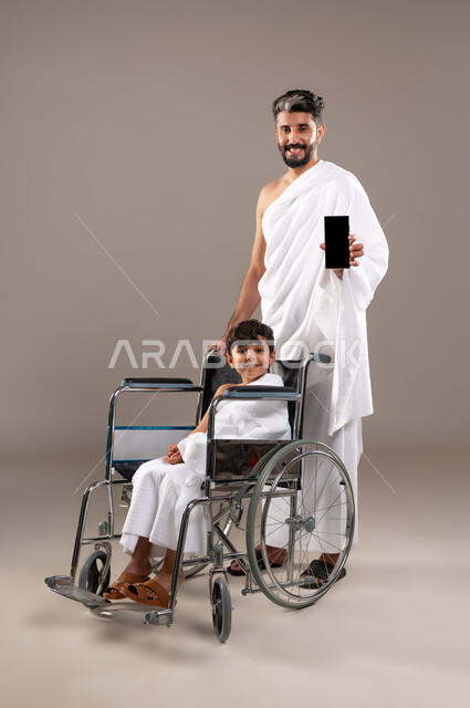 الاستعداد لاداء مناسك الحج والعمره والتقرب الى الله بالاعمال الصالحه، بورتريه لرجل عربي خليجي سعودي يرتدي ثوب الاحرام يجر ابنه الصبي المقعد من ذوي الاحتياجات الخاصه لاداء مناسك الحج او العمره، فريضه الحج هو الركن الخامس من أركان الاسلام لمن استطاع اليه سبيلا، خلفيه رماديه