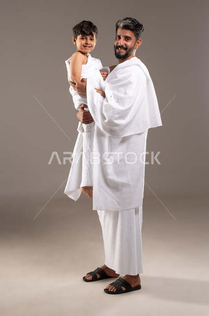 الاستعداد لاداء مناسك الحج والعمره والتقرب الى الله بالاعمال الصالحه، بورتريه لرجل عربي خليجي سعودي يرتدي ثوب الاحرام يحمل ابنه الصبي ويقوم بمداعبته بعد اداء الفريضه، الركن الخامس من أركان الاسلام لمن استطاع اليه سبيلا، اسلاميات وعباده، خلفيه رماديه
