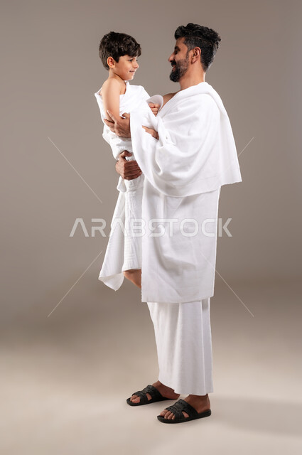 Preparing to perform the rituals of Hajj and Umrah and getting closer to God through good deeds, a portrait of a Saudi Gulf Arab man wearing the ihram dress carrying his boy son and caressing him after performing the obligatory prayers, the fifth pillar of Islam for those who can find a way to it, Islamic matters and worshipers, gray background