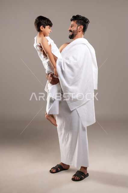 الاستعداد لاداء مناسك الحج والعمره والتقرب الى الله بالاعمال الصالحه، بورتريه لرجل عربي خليجي سعودي يرتدي ثوب الاحرام يحمل ابنه الصبي ويقوم بمداعبته بعد اداء الفريضه، الركن الخامس من أركان الاسلام لمن استطاع اليه سبيلا، اسلاميات وعباده، خلفيه رماديه