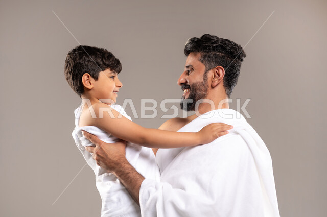 الاستعداد لاداء مناسك الحج والعمره والتقرب الى الله بالاعمال الصالحه، بورتريه لرجل عربي خليجي سعودي يرتدي ثوب الاحرام يحمل ابنه الصبي ويقوم بمداعبته بعد اداء الفريضه، الركن الخامس من أركان الاسلام لمن استطاع اليه سبيلا، اسلاميات وعباده، خلفيه رماديه