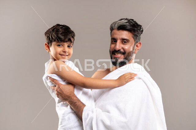الاستعداد لاداء مناسك الحج والعمره والتقرب الى الله بالاعمال الصالحه، بورتريه لرجل عربي خليجي سعودي يرتدي ثوب الاحرام يحمل ابنه الصبي ويقوم بمداعبته بعد اداء الفريضه، الركن الخامس من أركان الاسلام لمن استطاع اليه سبيلا، اسلاميات وعباده، خلفيه رماديه