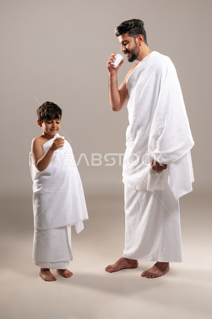 الاستعداد لاداء مناسك الحج والعمره والتقرب الى الله بالاعمال الصالحه، بورتريه لرجل وصبي عربي خليجي سعودي يرتديان ثوب الاحرام، الوقوف وشرب ماء زمزم بعد اداء الفريضه والنافله في مكه المكرمه، الركن الخامس من أركان الاسلام لمن استطاع اليه سبيلا، خلفيه رماديه