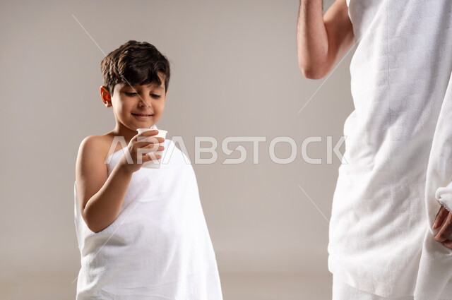 Preparing to perform the rituals of Hajj and Umrah and getting closer to God through good deeds, a portrait of a Saudi Arabian Gulf man and boy wearing the Ihram dress, standing and drinking Zamzam water after performing the obligatory and voluntary prayers in Mecca, the fifth pillar of Islam for those who have access to it, gray background
