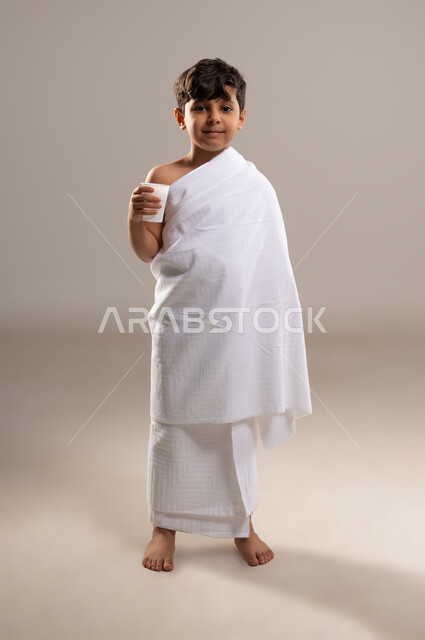 الاستعداد لاداء مناسك الحج والعمره والتقرب الى الله بالاعمال الصالحه، بورتريه لصبي عربي خليجي سعودي يرتدي ثوب الاحرام، الوقوف وشرب ماء زمزم بعد اداء الفريضه والنافله في مكه المكرمه، الركن الخامس من أركان الاسلام لمن استطاع اليه سبيلا، خلفيه رماديه