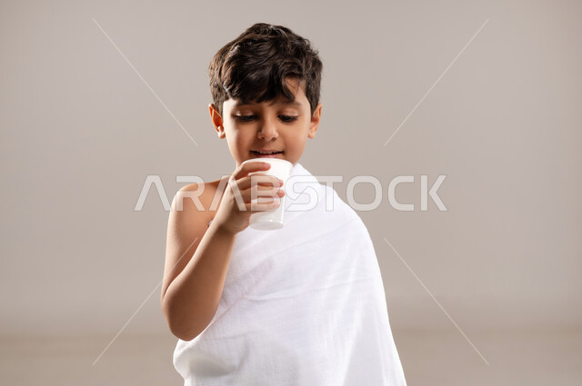 الاستعداد لاداء مناسك الحج والعمره والتقرب الى الله بالاعمال الصالحه، بورتريه لصبي عربي خليجي سعودي يرتدي ثوب الاحرام، الوقوف وشرب ماء زمزم بعد اداء الفريضه والنافله في مكه المكرمه، الركن الخامس من أركان الاسلام لمن استطاع اليه سبيلا، خلفيه رماديه