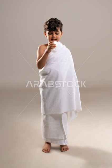 الاستعداد لاداء مناسك الحج والعمره والتقرب الى الله بالاعمال الصالحه، بورتريه لصبي عربي خليجي سعودي يرتدي ثوب الاحرام، الوقوف وشرب ماء زمزم بعد اداء الفريضه والنافله في مكه المكرمه، الركن الخامس من أركان الاسلام لمن استطاع اليه سبيلا، خلفيه رماديه