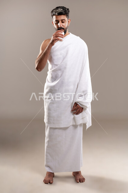 الاستعداد لاداء مناسك الحج والعمره والتقرب الى الله بالاعمال الصالحه، بورتريه لرجل عربي خليجي سعودي يرتدي ثوب الاحرام، الوقوف وشرب ماء زمزم بعد اداء الفريضه والنافله في مكه المكرمه، الركن الخامس من أركان الاسلام لمن استطاع اليه سبيلا، خلفيه رماديه