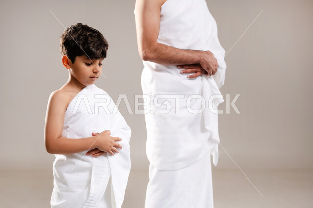 الاستعداد لاداء مناسك الحج والعمره والتقرب الى الله بالاعمال الصالحه، بورتريه لرجل وصبي عربيان خليجيان سعوديان يرتديان ثوب الاحرام، الوقوف وأداء فريضه الصلاه في المسجد الحرام في مكه المكرمه، الركن الثاني والخامس من أركان الاسلام، خلفيه رماديه