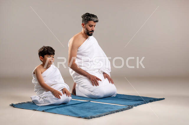 الاستعداد لاداء مناسك الحج والعمره والتقرب الى الله بالاعمال الصالحه، بورتريه لرجل وصبي عربيان خليجيان سعوديان يرتديان ثوب الاحرام يجلسان على سجاده الصلاه يؤديان فريضه الصلاه في المسجد الحرام في مكه المكرمه، أداء الركن الثاني والخامس من أركان الاسلام، خلفيه رماديه