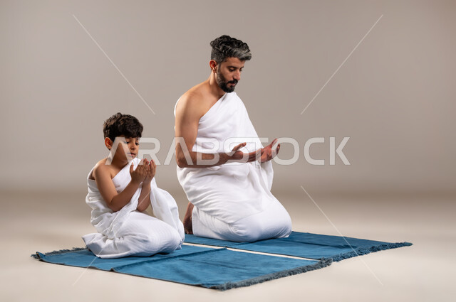 الاستعداد لاداء مناسك الحج والعمره والتقرب الى الله بالاعمال الصالحه، بورتريه لرجل وصبي عربيان خليجيان سعوديان يرتديان ثوب الاحرام يجلسان على سجاده الصلاه يرفعان يديهما للدعاء في المسجد الحرام في مكه المكرمه، أداء الركن الخامس من أركان الاسلام، خلفيه رماديه