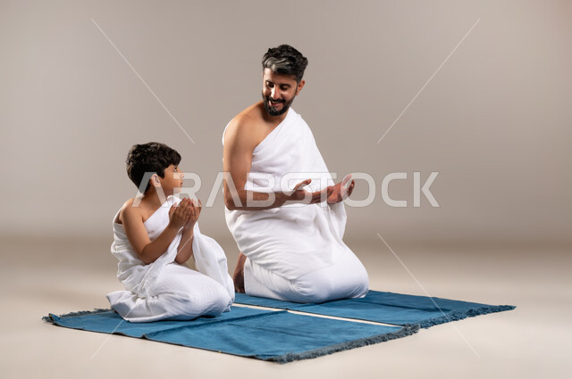 الاستعداد لاداء مناسك الحج والعمره والتقرب الى الله بالاعمال الصالحه، بورتريه لرجل وصبي عربيان خليجيان سعوديان يرتديان ثوب الاحرام يجلسان على سجاده الصلاه يرفعان يديهما للدعاء في المسجد الحرام في مكه المكرمه، أداء الركن الخامس من أركان الاسلام، خلفيه رماديه