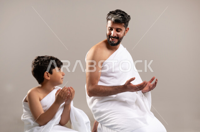 الاستعداد لاداء مناسك الحج والعمره والتقرب الى الله بالاعمال الصالحه، بورتريه لرجل وصبي عربيان خليجيان سعوديان يرتديان ثوب الاحرام يجلسان على سجاده الصلاه يرفعان يديهما للدعاء في المسجد الحرام في مكه المكرمه، أداء الركن الخامس من أركان الاسلام، خلفيه رماديه