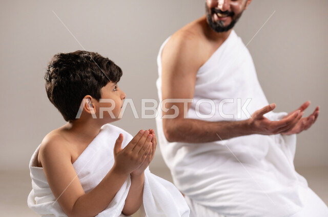 Preparing to perform the rituals of Hajj and Umrah and getting closer to God through good deeds, a portrait of a Saudi Gulf Arab man and boy wearing the Ihram dress, sitting on the prayer rug, raising their hands to pray in the Sacred Mosque in Mecca, performing the fifth pillar of Islam, gray background.