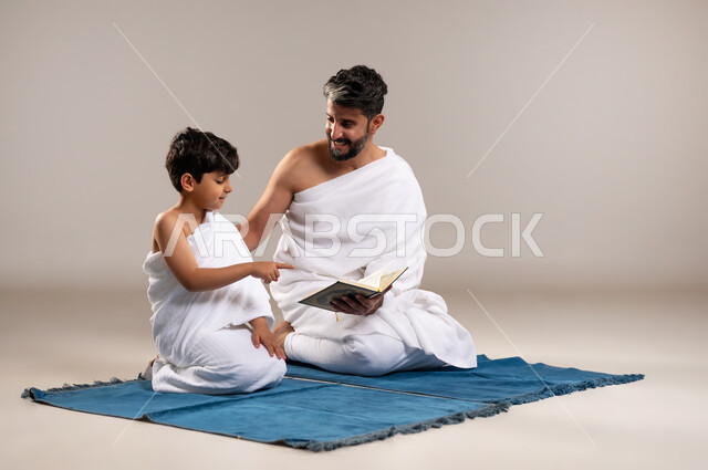 الاستعداد لاداء مناسك الحج والعمره والتقرب الى الله بالاعمال الصالحه، بورتريه لرجل وصبي عربيان خليجيان سعوديان يرتديان ثوب الاحرام يجلسان على سجاده الصلاه يقومان بقراءه المصحف الشريف في المسجد الحرام في مكه المكرمه، أداء الركن الخامس من أركان الاسلام، خلفيه رماديه