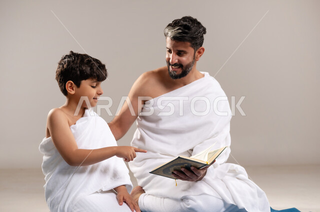 الاستعداد لاداء مناسك الحج والعمره والتقرب الى الله بالاعمال الصالحه، بورتريه لرجل وصبي عربيان خليجيان سعوديان يرتديان ثوب الاحرام يجلسان على سجاده الصلاه يقومان بقراءه المصحف الشريف في المسجد الحرام في مكه المكرمه، أداء الركن الخامس من أركان الاسلام، خلفيه رماديه