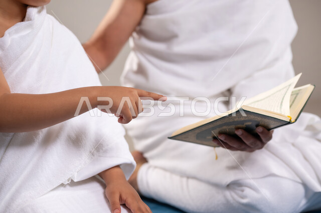 الاستعداد لاداء مناسك الحج والعمره والتقرب الى الله بالاعمال الصالحه، بورتريه لرجل وصبي عربيان خليجيان سعوديان يرتديان ثوب الاحرام يجلسان على سجاده الصلاه يقومان بقراءه المصحف الشريف في المسجد الحرام في مكه المكرمه، أداء الركن الخامس من أركان الاسلام، خلفيه رماديه