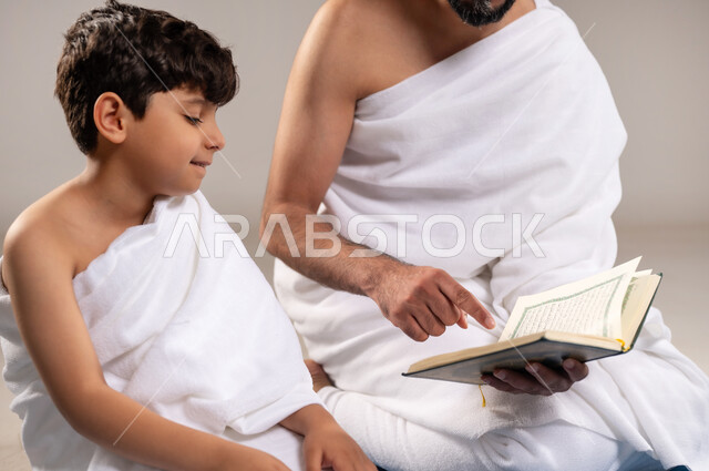 الاستعداد لاداء مناسك الحج والعمره والتقرب الى الله بالاعمال الصالحه، بورتريه لرجل وصبي عربيان خليجيان سعوديان يرتديان ثوب الاحرام يجلسان على سجاده الصلاه يقومان بقراءه المصحف الشريف في المسجد الحرام في مكه المكرمه، أداء الركن الخامس من أركان الاسلام، خلفيه رماديه
