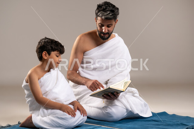 الاستعداد لاداء مناسك الحج والعمره والتقرب الى الله بالاعمال الصالحه، بورتريه لرجل وصبي عربيان خليجيان سعوديان يرتديان ثوب الاحرام يجلسان على سجاده الصلاه يقومان بقراءه المصحف الشريف في المسجد الحرام في مكه المكرمه، أداء الركن الخامس من أركان الاسلام، خلفيه رماديه