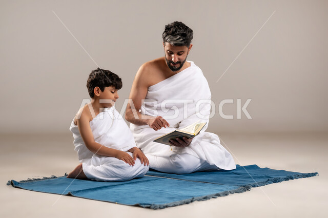 الاستعداد لاداء مناسك الحج والعمره والتقرب الى الله بالاعمال الصالحه، بورتريه لرجل وصبي عربيان خليجيان سعوديان يرتديان ثوب الاحرام يجلسان على سجاده الصلاه يقومان بقراءه المصحف الشريف في المسجد الحرام في مكه المكرمه، أداء الركن الخامس من أركان الاسلام، خلفيه رماديه