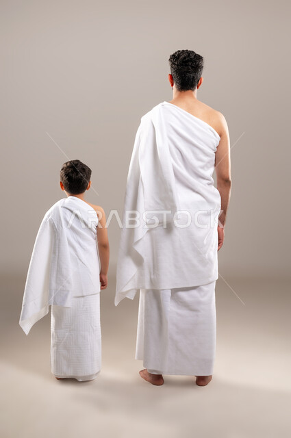 الاستعداد لاداء مناسك الحج والعمره والتقرب الى الله بالاعمال الصالحه، بورتريه بالطول الكامل لرجل وصبي عربيان خليجيان سعوديان يرتديان ثوب الاحرام استعدادا للذهاب الى المسجد الحرام في مكه المكرمه، أداء الركن الخامس من أركان الاسلام، خلفيه رماديه