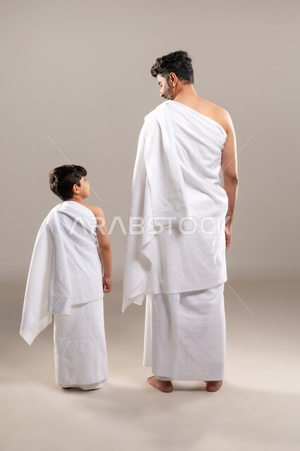 الاستعداد لاداء مناسك الحج والعمره والتقرب الى الله بالاعمال الصالحه، بورتريه بالطول الكامل لرجل وصبي عربيان خليجيان سعوديان يرتديان ثوب الاحرام استعدادا للذهاب الى المسجد الحرام في مكه المكرمه، أداء الركن الخامس من أركان الاسلام، خلفيه رماديه