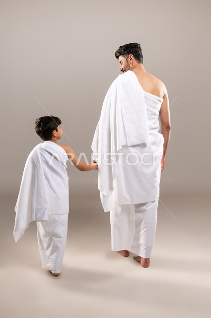 الاستعداد لاداء مناسك الحج والعمره والتقرب الى الله بالاعمال الصالحه، بورتريه بالطول الكامل لرجل وصبي عربيان خليجيان سعوديان يرتديان ثوب الاحرام استعدادا للذهاب الى المسجد الحرام في مكه المكرمه يمسكان بيد بعضهما، أداء الركن الخامس من أركان الاسلام، خلفيه رماديه