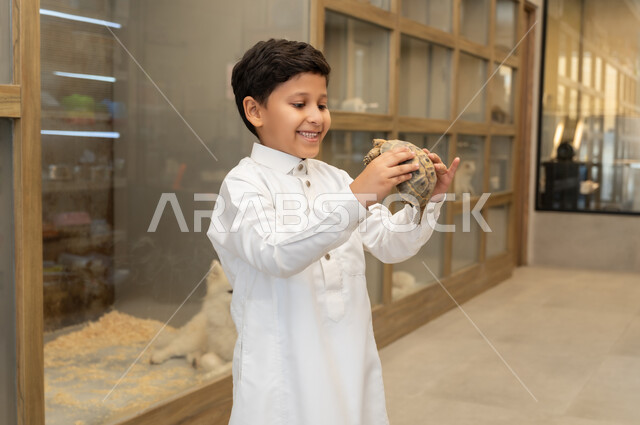 زيارة محلات ومتاجر بيع وشراء الحيوانات والطيور الأليفة، النظرالى السلحفاة اللطيفة بايماءات الفرح والسرور، أماكن خاصة لحفظ الحيوانات،  صورة مقربة لولدعربي خليجي سعودي  يرتدي الزي التقليدي يقوم بحمل سلحفاة وقضاء اوقات ممتعة ومسلية