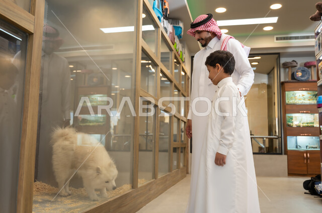 مفهوم الرفق بالحيوان الودود المخلص، متاجر بيع وشراء الحيوانات الأليفة، رجل عربي خليجي سعودي مبتسم يرتدي الثوب التقليدي والشماغ  يجلس برفقة ابنه امام نافذة زجاجية ، ايماءات السعادة والسروربمشاهدة الكلب الابيض الوفي