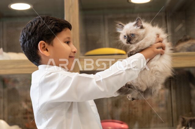 تربية ورعاية القطط في المنازل، مفهوم الرفق بالحيوان والعطف عليه، ولد عربي خليجي سعودي مبتسم يرتدي الثوب التقليدي يحمل بيده قطة اليفة، محلات لبيع الحيوانات الاليفة، قضاء وقت ممتع في اللعب والمرح مع القط المدلل