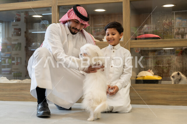 متاجربيع وشراء حاجات مستلزمات الحيوانات الاليفة، مفهوم الرفق بالحيوان الودود المخلص، رجل عربي خليجي سعودي مبتسم يرتدي الثوب التقليدي يجلس مع ولده، ايماءات السعادة والمرح واللعب مع الكلب الابيض الوفي، قضاء وقت ممتع في اللعب مع الحيوان الاليف
