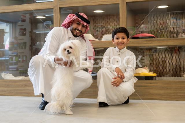 مفهوم الرفق بالحيوان الودود المخلص، رجل عربي خليجي سعودي مبتسم يرتدي الثوب التقليدي يجلس مع ولده، متاجربيع وشراء حاجات مستلزمات الحيوانات الاليفة، ايماءات السعادة والمرح واللعب مع الكلب الابيض الوفي، قضاء وقت ممتع في اللعب مع الحيوان الاليف