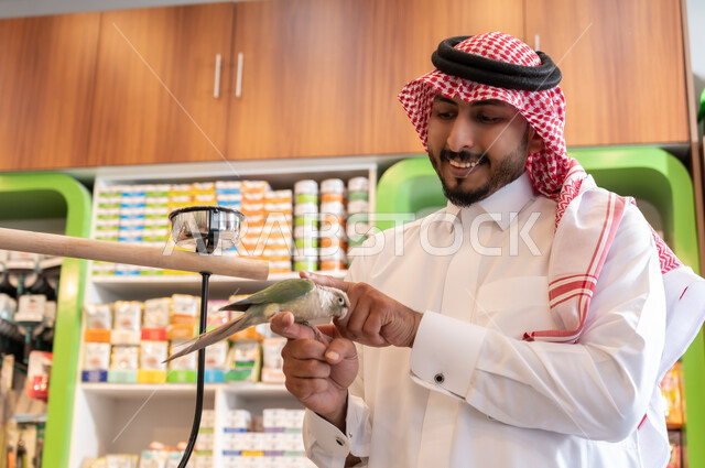 أماكن خاصة لحفظ العصافير، زيارة محلات ومتاجر بيع وشراء الحيوانات والطيور الأليفة، الشغف بتربية الحيوانات الاليفة في المنزل، صورة مقربة لولد عربي خليجي سعودي يرتدي الثوب التقليدي يضع عصفور ملون على رأسه، قضاء اوقات ممتعة ومسلية، ينظر الى الكاميرا بايماءات الفرح والسرور