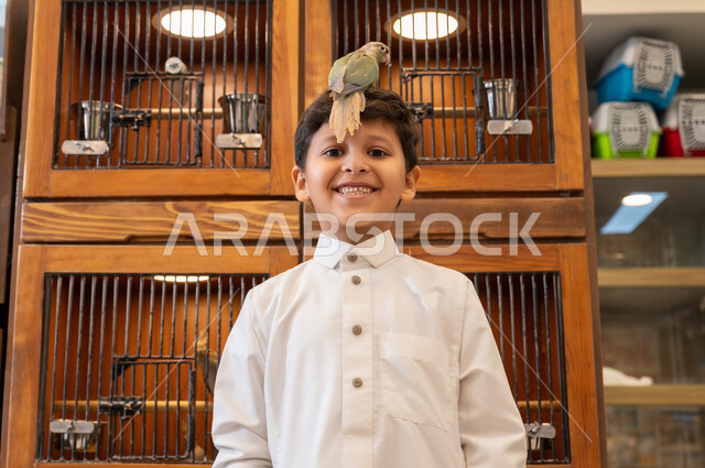 أماكن خاصة لحفظ العصافير، زيارة محلات ومتاجر بيع وشراء الحيوانات والطيور الأليفة، الشغف بتربية الحيوانات الاليفة في المنزل، صورة مقربة لولد عربي خليجي سعودي يرتدي الثوب التقليدي يضع عصفور ملون على رأسه، قضاء اوقات ممتعة ومسلية، ينظر الى الكاميرا بايماءات الفرح والسرور