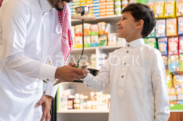 زيارة المحلات الخاصة بشراء الحيوانات والطيور الاليفة، الشغف بتربية الحيوانات الاليفة في المنزل، النظرالى العصفور، العناية بصحة الطيور وتوفيرالاغذية المناسبة لهم،  صورة مقربة لرجل عربي خليجي سعودي يرتدي الثوب التقليدي والشماغ برفقة ولده يحمل بيده طائر ملون