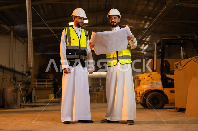 الاشراف على سير خطة العمل، حرف ومهن شعبية، حوارات ونقاشات هادفة، معمل تصنيع الأثاث الخشبي، ورشات النجارة في السعودية، مهندسان عربيان خليجيان سعوديان يرتديان سترة وخوذة الحماية يحملان مخططات هندسية