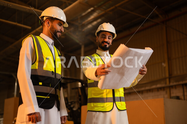 الاشراف على سير خطة العمل، حرف ومهن شعبية، حوارات ونقاشات هادفة، معمل تصنيع الأثاث الخشبي، ورشات النجارة في السعودية، مهندسان عربيان خليجيان سعوديان يرتديان سترة وخوذة الحماية يحملان مخططات هندسية