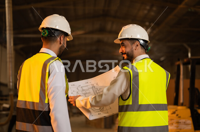 حرف ومهن شعبية، حوارات ونقاشات هادفة، معمل تصنيع الأثاث الخشبي، ورشات النجارة في السعودية، صورة مقربة من الخلف لمهندسان عربيان خليجيان سعوديان يرتديان سترة وخوذة الحماية يحملان مخططات هندسية، الاشراف على سير خطة العمل