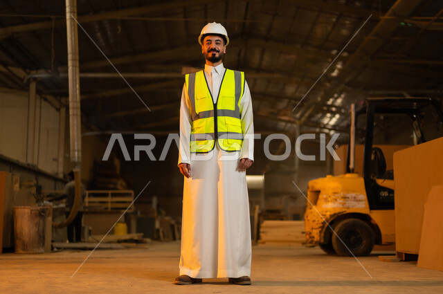 الوقوف باستقامة والنظر إلى الكاميرا بإيماءات الثقة بالنفس والجدية, ورشات عمل ومهن تقليدية قديمة, مصانع ومعامل في المملكة العربية السعودية, مهندس عربي خليجي سعودي يرتدي الخوذة وسترة الحماية يعمل في معمل صناعة الخشب, الاشراف على سير خطة العمل