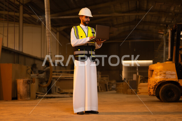 التحكم بالمعدات باستخدام التقنية والتكنولوجيا، معمل تصنيع الأثاث الخشبي، مهن ووظائف صناعية، ورشات النجارة في السعودية، المتابعة والإشراف على آلات المعمل، مهندس عربي خليجي سعودي يرتدي سترة وخوذة الحماية يحمل بيده الحاسوب المحمول