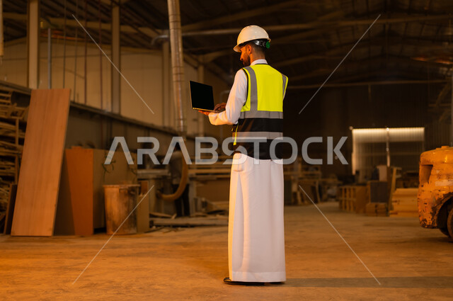 Industrial jobs and professions, carpentry workshops in Saudi Arabia, controlling equipment using technology and technology, monitoring and supervising factory machines, wooden furniture manufacturing factory, a picture from the back of a Saudi Arabian Gulf engineer wearing a protective jacket and helmet holding a laptop in his hand