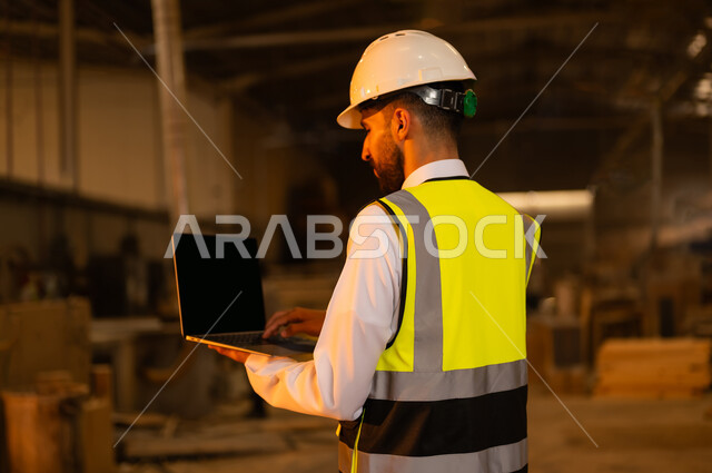 ورشات النجارة في السعودية، التحكم بالمعدات باستخدام التقنية والتكنولوجيا، المتابعة والإشراف على آلات المعمل، معمل تصنيع الأثاث الخشبي، مهن ووظائف صناعية، صورة مقربة من الخلف لمهندس عربي خليجي سعودي يرتدي سترة وخوذة الحماية يحمل بيده الحاسوب المحمول