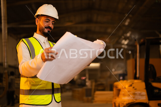 الاشراف على سير خطة العمل، حرف ومهن شعبية، حوارات ونقاشات هادفة، معمل تصنيع الأثاث الخشبي، ورشات النجارة في السعودية، صورة مقربة لمهندس عربي خليجي سعودي يرتدي سترة وخوذة الحماية يحمل بيده مخططات هندسية