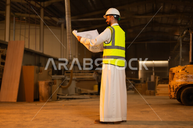 حرف ومهن شعبية، الاشراف على سير خطة العمل، حوارات ونقاشات هادفة، معمل تصنيع الأثاث الخشبي، ورشات النجارة في السعودية، صورة من الخلف لمهندس عربي خليجي سعودي يرتدي سترة وخوذة الحماية يحمل بيده مخططات هندسية