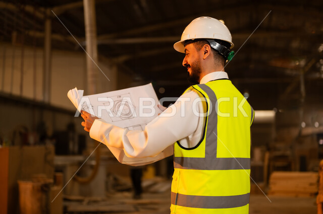 حرف ومهن شعبية، الاشراف على سير خطة العمل، حوارات ونقاشات هادفة، معمل تصنيع الأثاث الخشبي، ورشات النجارة في السعودية، صورة مقربة من الخلف لمهندس عربي خليجي سعودي يرتدي سترة وخوذة الحماية يحمل بيده مخططات هندسية