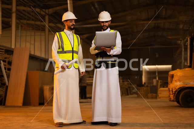 التحكم بالمعدات باستخدام التقنية والتكنولوجيا، معمل تصنيع الأثاث الخشبي، مهن ووظائف صناعية، ورشات النجارة في السعودية، المتابعة والإشراف على آلات المعمل، مهندس عربي خليجي سعودي يرتدي سترة وخوذة الحماية يحمل بيده الحاسوب المحمول يقف برفقة زميله