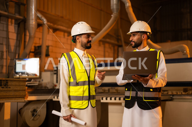 مهن ووظائف صناعية، ورشات النجارة في السعودية، معمل تصنيع الأثاث الخشبي، التحكم بالمعدات باستخدام التقنية والتكنولوجيا، المتابعة والإشراف على آلات المعمل، مهندس عربي خليجي سعودي يرتدي سترة وخوذة الحماية يحمل بيده الحاسوب المحمول يقف برفقة زميله